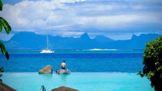 A boy sitting on a rock in a pool overlooking the blue waters of Tahiti.