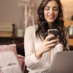 Woman Sitting on Sofa While Looking at Phone With Laptop on Lap