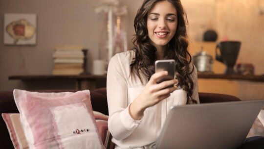 Woman Sitting on Sofa While Looking at Phone With Laptop on Lap